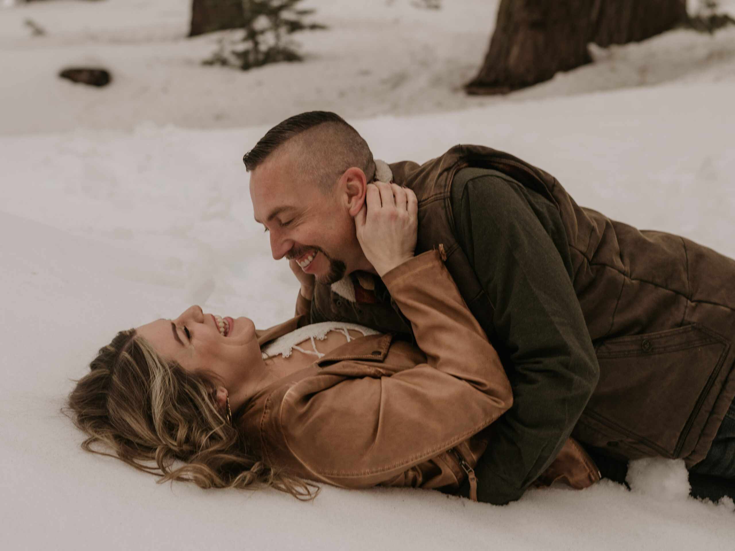 Couples Photoshoot or couple laying in the snow and laughing during their session. Snowy mountains and beautiful big trees!