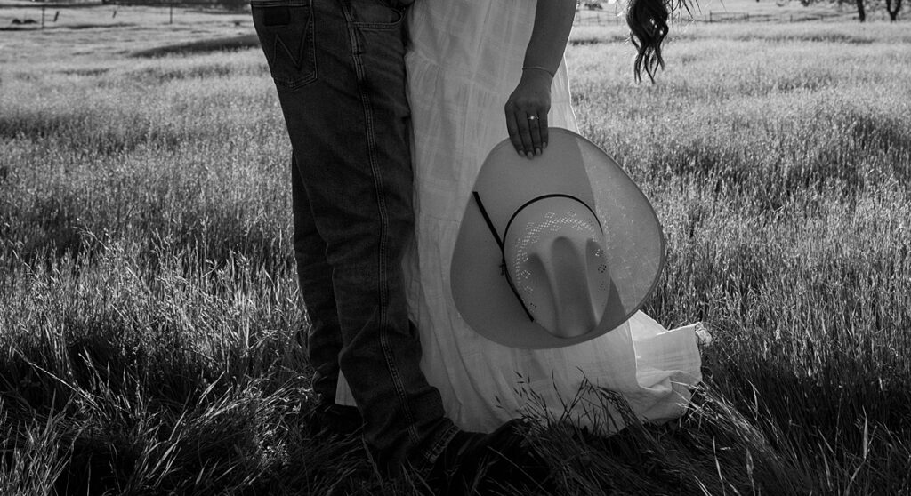 A b+w photo shows a woman holding her fiancé's cowboy hat with her left hand, showcasing the ring during engagement photos in California 
