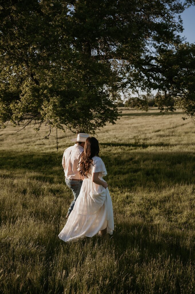 A young couple hold hands at golden hour as they stroll through their farm for engagement photos california