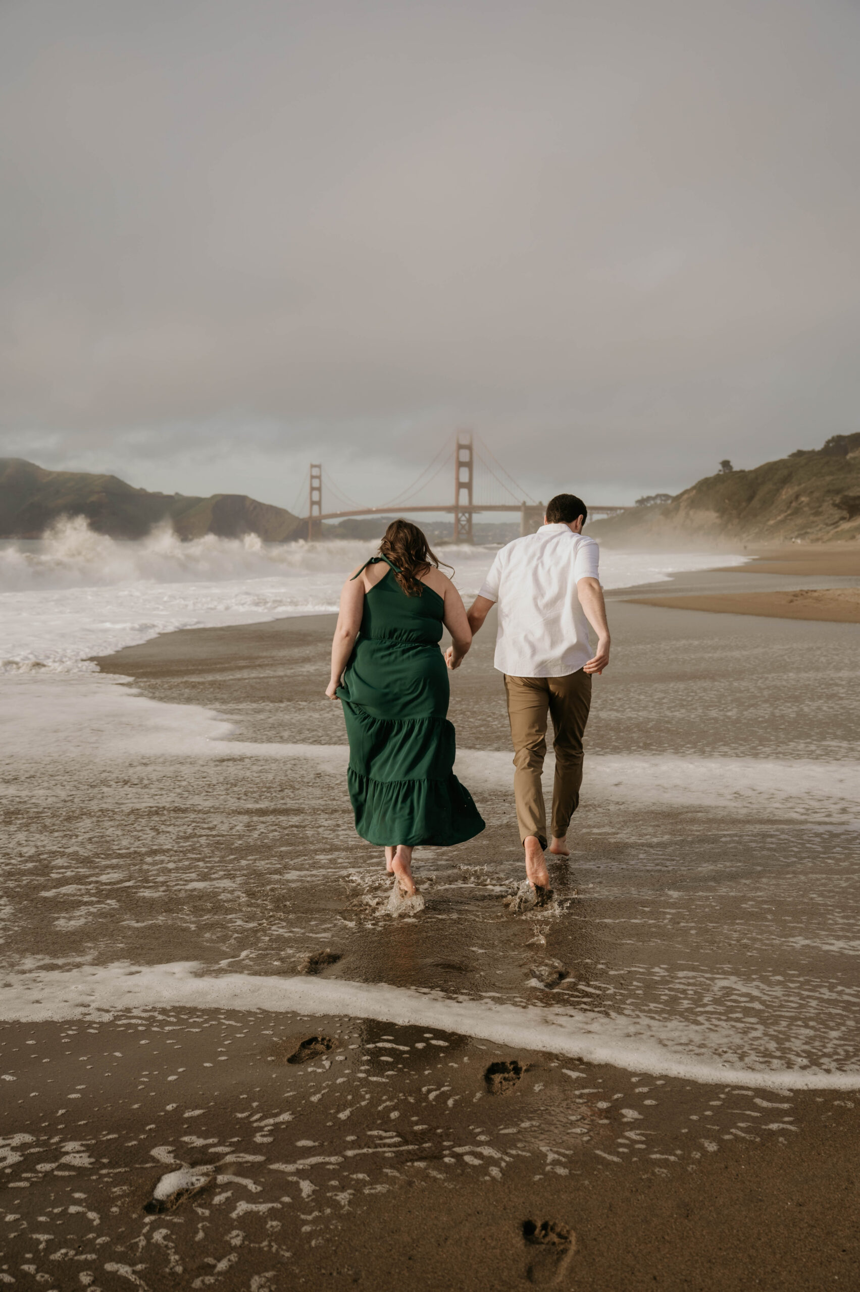 couple holding hands on the beach with golden gate bridge in front of them during their engagement photos
