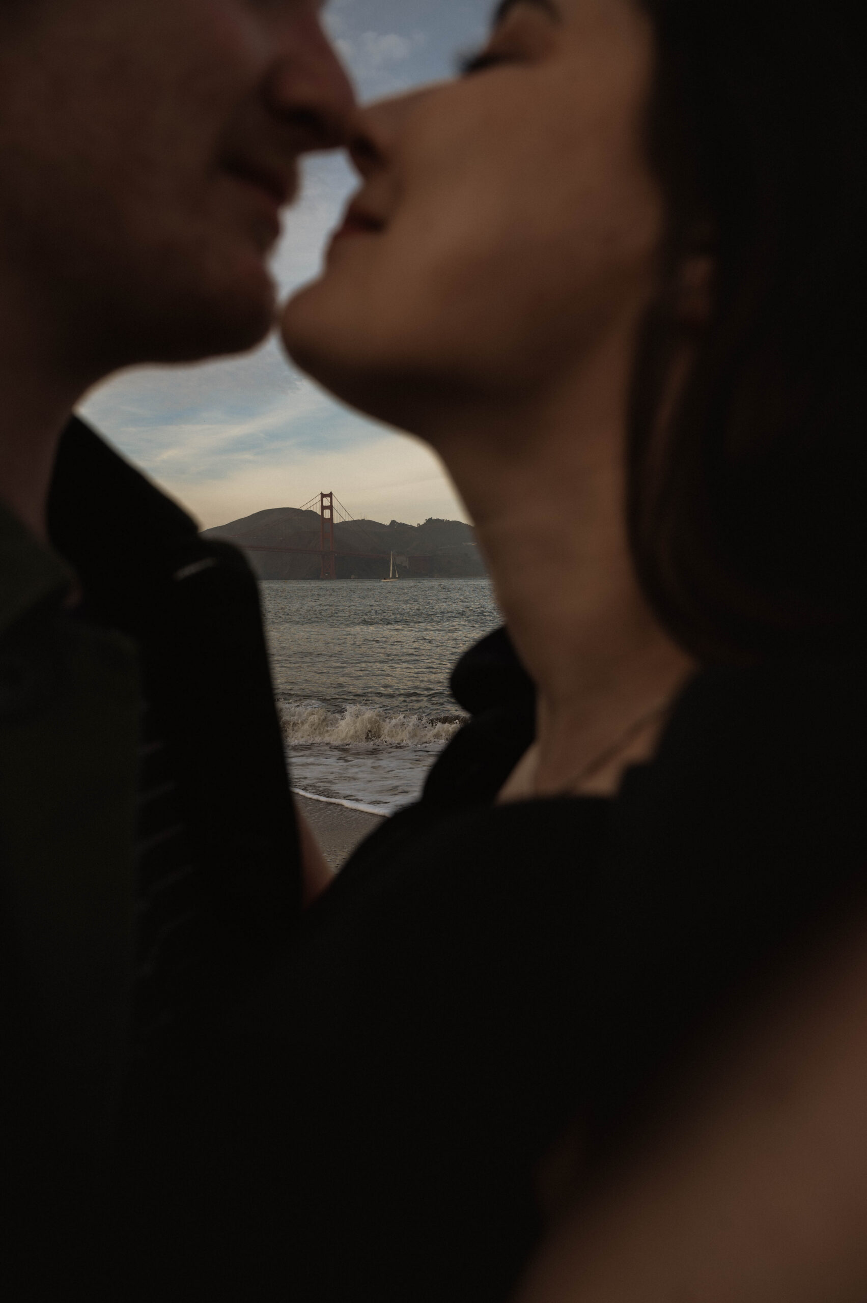 a romantic moment between an engaged couple during their engagement photos, where they embrace with the crashing waves and the Golden Gate Bridge behind them