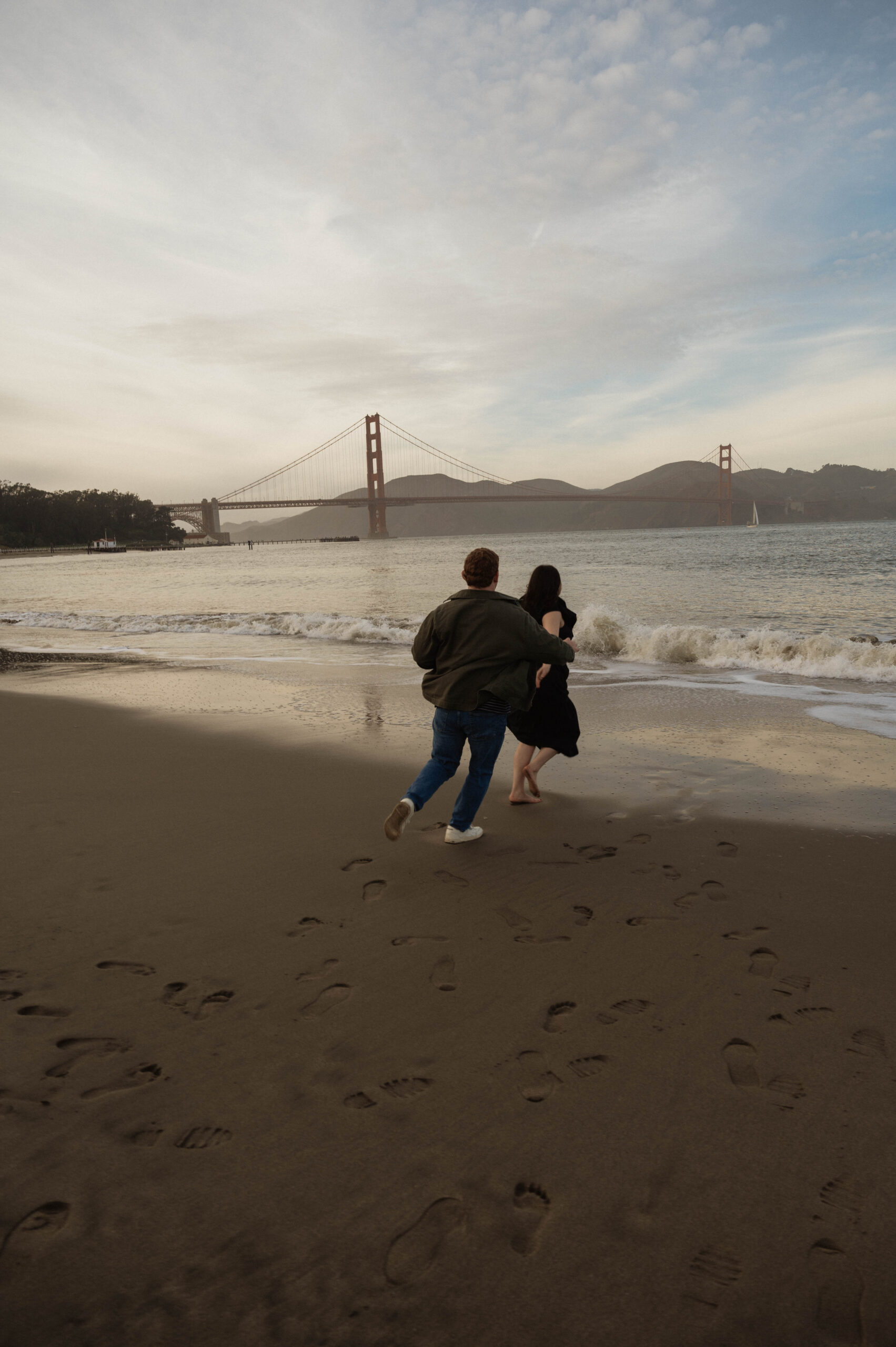 A couple celebrates their engagement by playfully chasing each other on the beach of San Francisco during their engagement photography session.