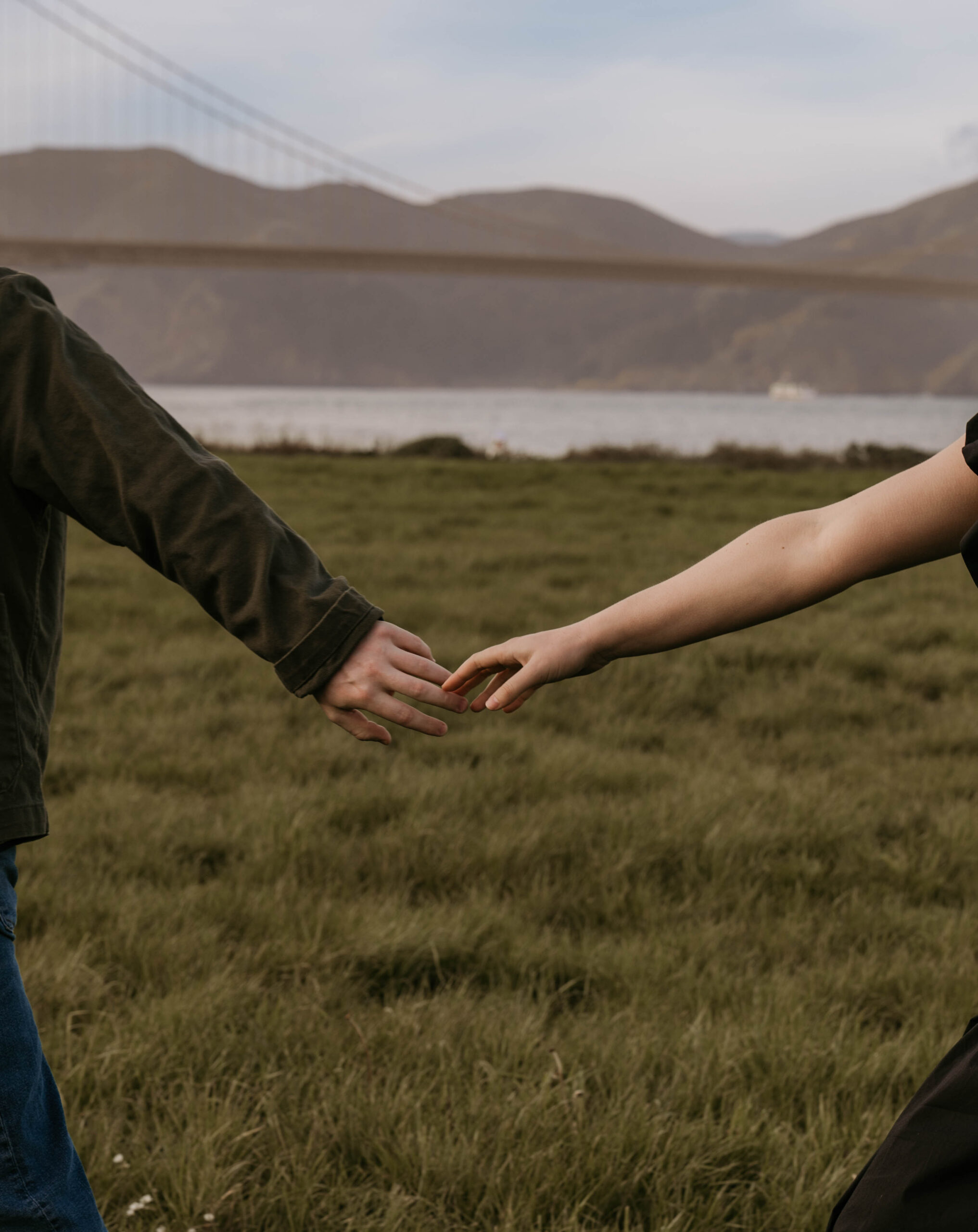 newly engaged couple reaches for each other's hands on the lush grass of Crissy Field in San Francisco